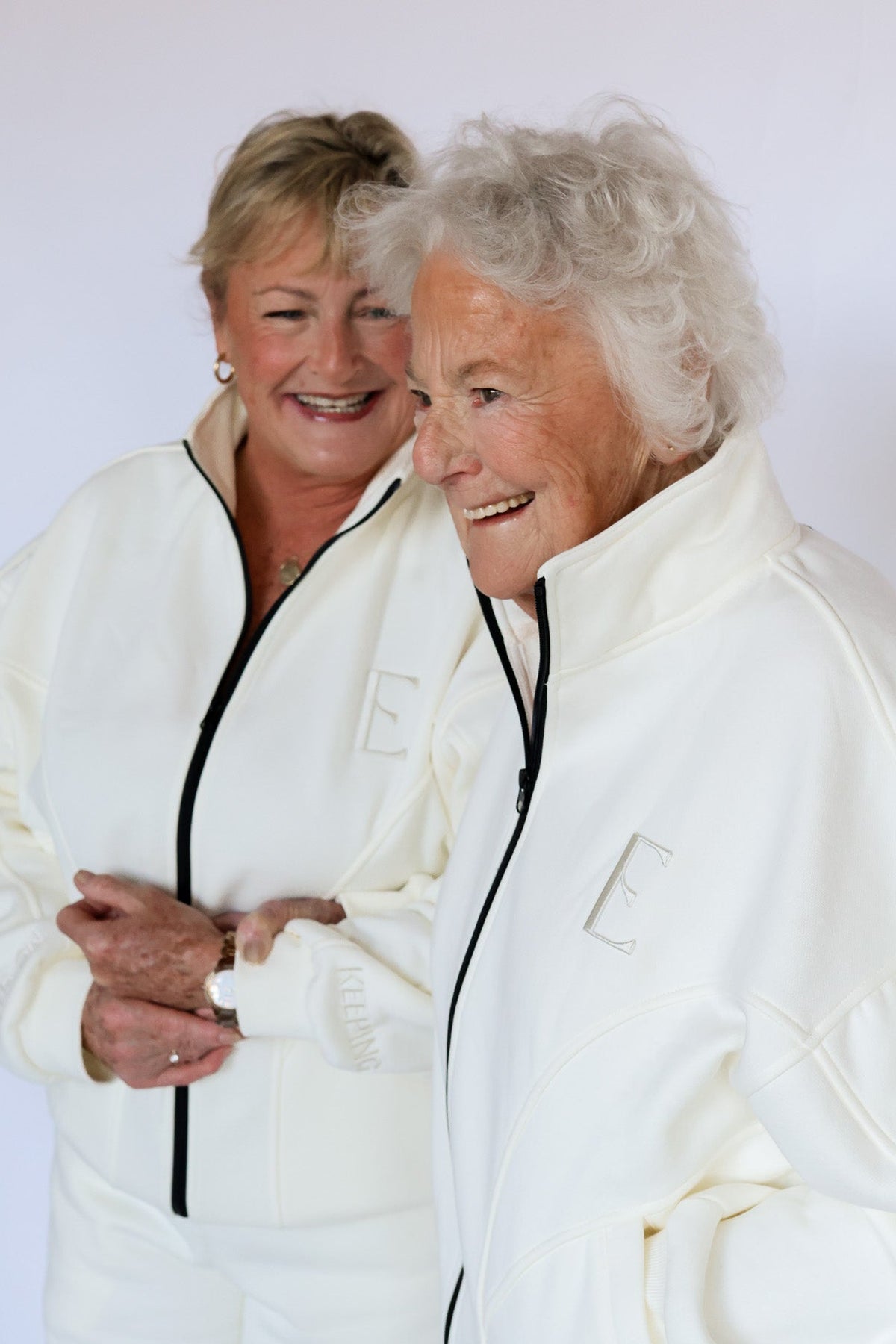 Two smiling women wearing matching white zip-up tracksuits with black zippers and a large embroidered letter 'E' on the chest, posed against a plain light background.