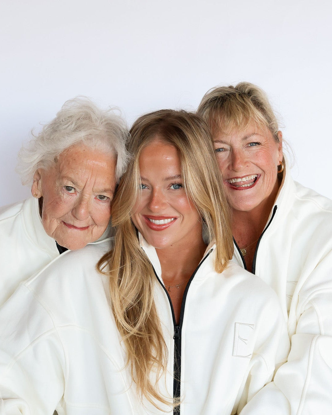 Three women wearing matching white zip-up jackets pose together against a plain white background.