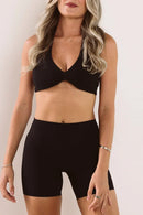 A woman wearing a black two-piece activewear set consisting of a halter-style sports bra with a front twist detail and matching high-waisted biker shorts, shown against a plain light background.