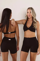 Two women modeling black athletic wear against a plain light background, showing front and back views of a matching sports bra and biker shorts set.