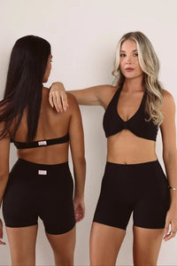 Two women modeling black athletic wear against a plain light background, showing front and back views of a matching sports bra and biker shorts set.