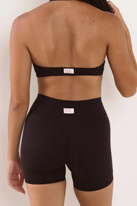 Rear view of a model wearing a black strapless bandeau top and matching high-waisted black shapewear shorts against a plain off-white background.