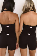 Two women shown from the back wearing matching black strapless biker shorts with a small white brand tag at the waistband, standing against a plain light background.