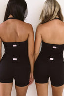 Two women shown from the back wearing matching black halter singlet and shorts with ELE brand tag at the center back, standing against a plain off-white wall.