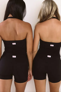 Two women shown from the back wearing matching black halter singlet and shorts with ELE brand tag at the center back, standing against a plain off-white wall.
